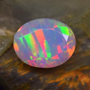May include: A round, faceted opal gemstone with a play of color. The opal displays flashes of red, green, and orange hues within a translucent white base. The gemstone is set against a natural, textured background.