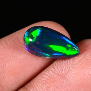 Puede incluir: Una piedra preciosa de ópalo negro en forma de pera que presenta un color base oscuro con destellos iridiscentes vibrantes de verde neón y azul eléctrico en su superficie pulida y lisa, mostrando un juego de colores distintivo en la piedra.