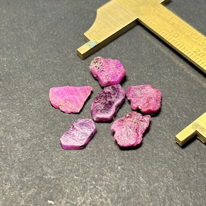 Peut inclure: Six pierres précieuses de rubis brut de différentes tailles et formes. Les pierres précieuses sont disposées en grappe sur une surface gris foncé. Une règle est visible en arrière-plan.