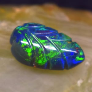 May include: A carved dark blue and green opal gemstone shaped like a leaf. The surface of the stone features deep grooves and a polished finish, displaying vibrant flashes of neon green and deep blue color play throughout the entire piece.