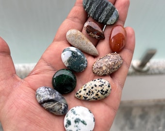 Peut inclure: Une collection de pierres précieuses polies de différentes formes et couleurs, dont des motifs verts, bruns, blancs et mouchetés. Les pierres sont tenues dans une main, mettant en valeur leurs textures naturelles et leurs designs uniques. Idéal pour la joaillerie ou la collection.