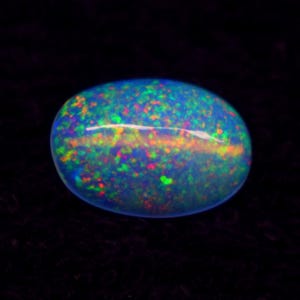 May include: Oval-shaped opal gemstone with a dark blue base color. The stone displays a vibrant play-of-color with flashes of red, green, and yellow. The opal is set against a black background, highlighting its iridescent qualities.