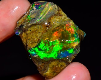 Puede incluir: Una piedra de ópalo en bruto, sin tallar, con colores vibrantes. La piedra muestra destellos de verde, naranja, azul y amarillo, sobre una matriz marrón. El ópalo se sostiene entre los dedos, mostrando su belleza natural.
