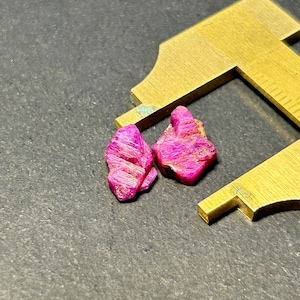 May include: Two pink ruby crystals, each with a rough, faceted surface, are measured with a caliper. The crystals are a deep pink colour and have a natural, unpolished appearance.