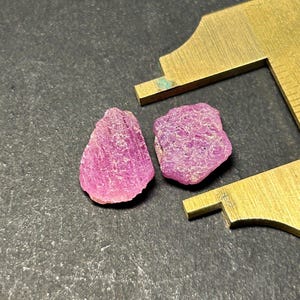 Könnte beinhalten: Zwei rohe rosa Rubine auf einer grauen Oberfläche. Die Steine sind etwa 1 cm groß.