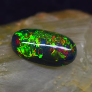 May include: A polished, oval-shaped black opal gemstone. The opal displays vibrant internal fire with flashes of green, red, and yellow. The gemstone is set against a neutral, textured background, highlighting its natural beauty.