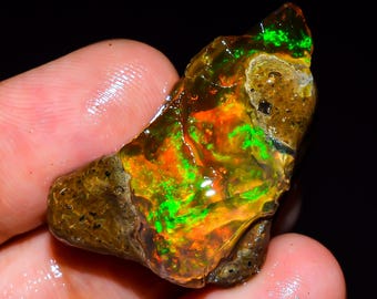 May include: A rough-cut opal gemstone with vibrant colors. The stone displays a mix of orange, green, and brown hues. The opal is held in a hand, showcasing its natural, irregular shape and unique color play. The gemstone is a natural mineral.