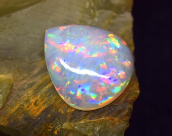 May include: A polished, pear-shaped opal gemstone with a milky white base. The opal displays vibrant flashes of color, including red, blue, green, and yellow, across its surface. The gemstone is set against a rough, textured, green and brown stone.