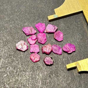 May include: A collection of pink ruby gemstones, some with a hexagonal shape, on a gray surface.