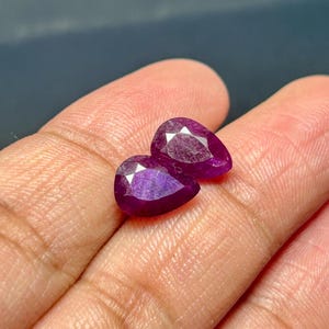 Könnte beinhalten: Zwei birnenförmige Edelsteine, beide in einem tiefen Lila, werden auf einer Hand gezeigt. Die Edelsteine sind facettiert und haben einen leichten Schimmer.
