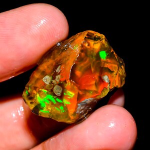 May include: A raw, unpolished gemstone featuring a translucent orange and amber base with vibrant, iridescent flashes of neon green and yellow light. The stone has an irregular, jagged shape with visible natural inclusions and a glossy surface.