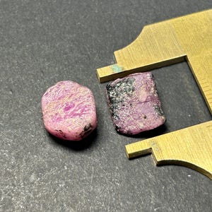 Può includere: Due pietre preziose grezze di colore rosa e nero. Le pietre preziose hanno una forma irregolare e una finitura grezza e naturale.