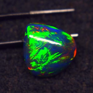 Puede incluir: Una piedra preciosa de ópalo negro pulido con vibrantes destellos verdes, azules y rojos. La gema está sujeta por unas pinzas, mostrando su juego de colores único y su belleza natural. La superficie del ópalo es lisa y reflectante.