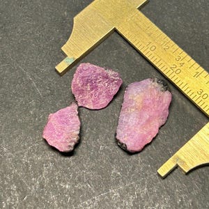 Könnte beinhalten: Drei rosafarbene Rubinkristalle mit schwarzen Einschlüssen, gemessen mit einem Lineal in Zentimetern.