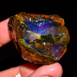 May include: A rough opal gemstone with a brown, textured exterior and a vibrant interior. The interior displays a play of color with blues, greens, and hints of red and yellow. The gemstone is held in a hand against a black background.