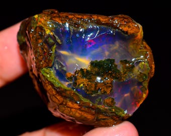 May include: A rough opal gemstone with a brown, textured exterior and a vibrant interior. The interior displays a play of color with blues, greens, and hints of red and yellow. The gemstone is held in a hand against a black background.