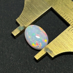 Könnte beinhalten: Ein weißer ovaler Opal-Edelstein mit irisierenden Schimmern in Grün, Rot und Orange. Der Opal wird mit einer Schieblehre gemessen.
