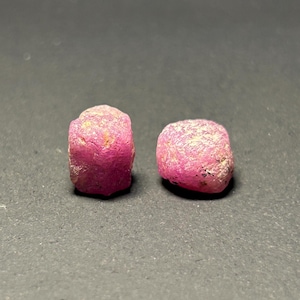 May include: Two pink ruby gemstones, each with a rough, faceted surface. The gemstones are a deep pink color and have a slightly translucent appearance.