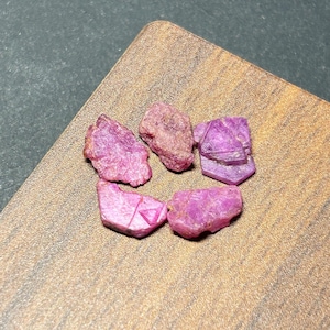 May include: Five rough, uncut ruby gemstones in varying shades of pink and red are arranged on a wooden surface. The gemstones have a natural, irregular shape and texture, with visible crystalline structures. The wooden surface has a warm, brown tone.