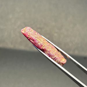 May include: A close-up of a pink ruby crystal with gold inclusions, held by tweezers.