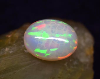 May include: A round, polished opal gemstone with a white base color and internal play-of-color. The opal displays flashes of green, red, and pink hues. The gemstone is set against a rough, tan-colored rock.