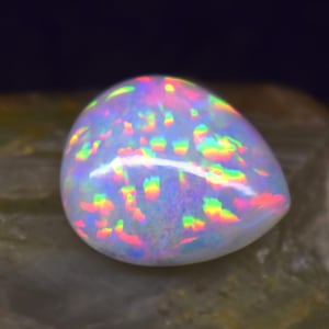 Könnte beinhalten: Ein birnenförmiger, schillernder Opal-Edelstein. Der Opal zeigt eine milchig-weiße Grundfarbe mit Regenbogenfarben, darunter Rot, Grün und Blau. Der Edelstein ist vor einem neutralen Hintergrund platziert.
