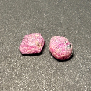 May include: Two pink ruby gemstones, each with a rough, irregular shape. The rubies have a deep pink color and are set against a dark gray background.
