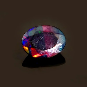 Puede incluir: Una piedra preciosa ovalada de color oscuro con un brillo iridiscente multicolor. La piedra tiene un patrón de color azul oscuro, morado, rojo y verde.
