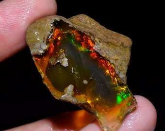 May include: A rough-cut opal gemstone with vibrant internal colors of red, orange, and green. The opal is held in a hand, showcasing its natural, unpolished texture and earthy tones. The stone's surface displays a play of light.