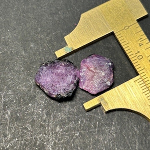 May include: Two purple rough cut gemstones, likely rubies, are measured with a ruler. The ruler is marked in millimetres.