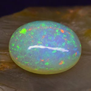Könnte beinhalten: Ein polierter, ovaler Opal-Edelstein mit milchig-weißer Basis. Der Stein zeigt lebhafte Blitze in Grün, Orange und Blau, mit einem markanten weißen Streifen. Der Opal liegt vor einem neutralen Hintergrund.