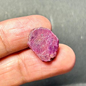 Puede incluir: Primer plano de una piedra preciosa de rubí rojo intenso con un acabado rugoso y natural. El rubí se sostiene en una mano, mostrando su tamaño y color.