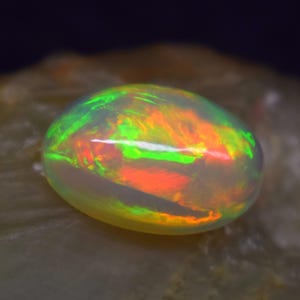 Puede incluir: Una vibrante piedra preciosa de ópalo de forma ovalada. El ópalo muestra un juego de colores con destellos verdes, naranjas y amarillos. La gema se encuentra sobre un fondo neutro y texturizado, resaltando sus cualidades iridiscentes.