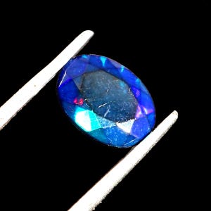 May include: A blue oval-shaped opal gemstone with iridescent flashes of color. The gemstone is held in a pair of tweezers against a black background.
