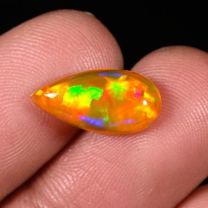 Peut inclure: Une pierre d'opale en forme de goutte d'eau. L'opale présente une couleur de base orange vif avec des éclairs internes de vert, de bleu et de rose, créant un effet de jeu de couleurs. La pierre est tenue entre les doigts sur fond sombre.