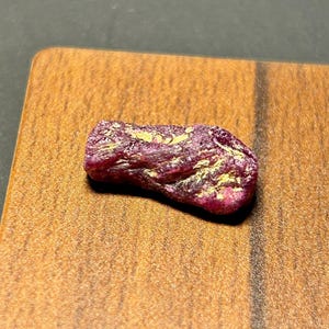 Può includere: Una pietra preziosa di rubino rosa grezza con inclusioni dorate. Il rubino ha una forma irregolare e una finitura naturale non lucidata.