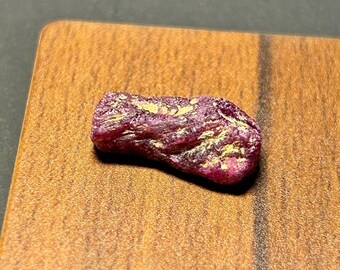 May include: A rough, pink ruby gemstone with gold inclusions. The ruby is irregularly shaped and has a natural, unpolished finish.