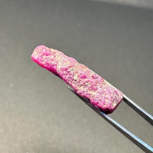 May include: A close-up of a pink ruby gemstone with a rough, natural surface. The ruby is held by tweezers and has a slightly elongated shape.