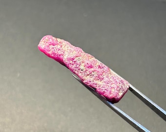 May include: A close-up of a pink ruby gemstone with a rough, natural surface. The ruby is held by tweezers and has a slightly elongated shape.