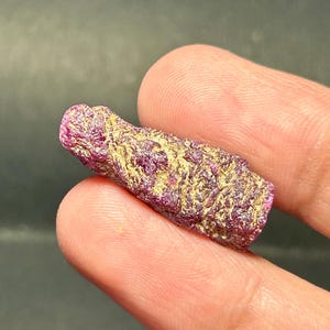 Może przedstawiać: Surowy, różowy kryształ rubinu z złotym połyskiem. Kryształ jest wydłużony i ma spiczasty koniec.