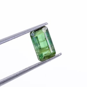 May include: A rectangular, transparent, green gemstone is held by silver tweezers against a white background. The gemstone has visible internal structures and a polished surface, showcasing its clarity and color. The gem is a vibrant shade of green.