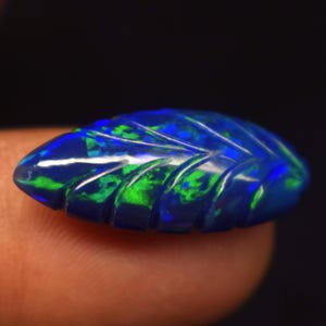 May include: A carved dark blue opal gemstone shaped like a leaf, featuring vibrant green and bright blue flashes of color throughout the surface. The stone has a polished finish with deep grooves that define the leaf vein pattern across the top.
