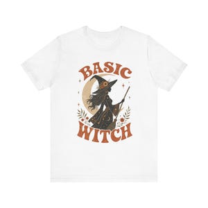 May include: A white short-sleeve t-shirt featuring a graphic print of a witch holding a broomstick in front of a crescent moon. The text BASIC WITCH is printed in a curved, textured orange font above and below the central illustration.