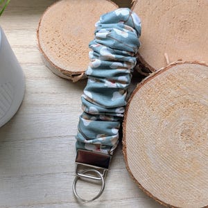 May include: A fabric keychain featuring a gathered, ruffled design in a dusty blue color with a white floral pattern. The fabric is attached to a silver-toned metal hardware clamp that connects to a circular metal split ring for holding keys.