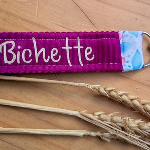 Puede incluir: Un llavero de tela magenta con el nombre "Bichette" en escritura blanca. El llavero tiene un cierre plateado y un adorno de tela estampada. Espigas de trigo en primer plano.