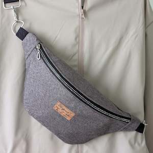 May include: A grey fanny pack with a black zip and strap. The bag features a rectangular patch with the text "Lil good!" on it. The bag is made of a textured fabric and has a silver-coloured clasp.