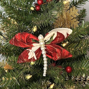 May include: A red and white fabric dragonfly ornament with a pearl and rhinestone embellishment. The ornament is hanging on a green Christmas tree.