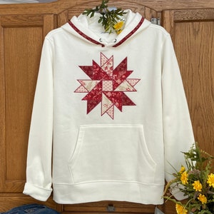 Peut inclure: Un sweat à capuche blanc avec un motif d'étoile en patchwork rouge et blanc sur le devant. Le sweat à capuche a un cordon de serrage rouge et une poche kangourou.