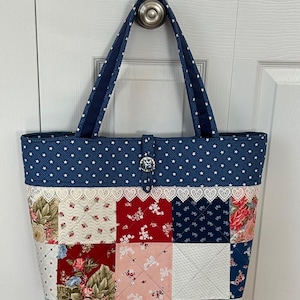 Op de afbeelding: Een blauwe en witte patchwork tote bag met een witte kantversiering en een sterrenpatroon. De tas heeft een blauwe band en een zilveren knoopsluiting.