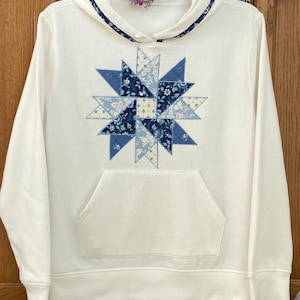 May include: A white hooded sweatshirt with a blue and white patchwork star design on the front. The sweatshirt has a kangaroo pocket and a drawstring hood.